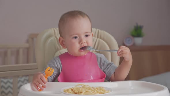 Toddler eats spaghetti for dinner by himself holding two forks in his hands