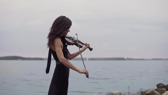Beautiful Girl in Black Dress Plays Violin at Sea Background. Art Concept in 