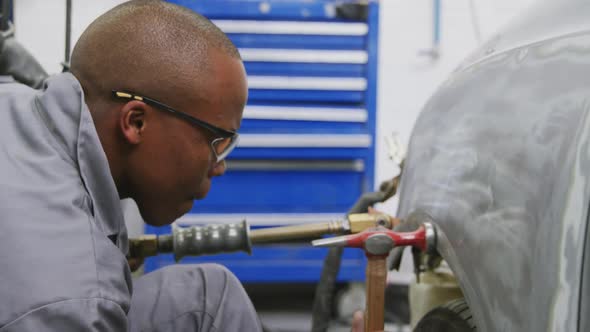 African American male car mechanic holding a screwdriver and using a hammer to repair a car