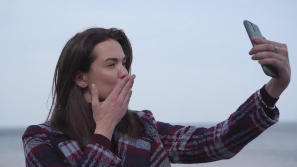 Close-up of Cheerful Brunette Girl Sending Air Kiss at Selfie Camera. Young Positive Caucasian Woman