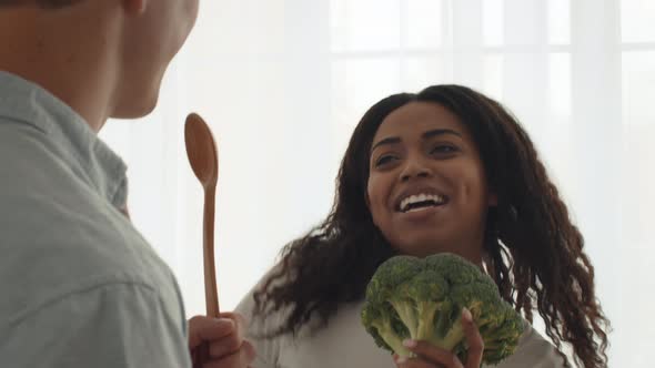 Cheerful Diverse Spouses Singing Holding Broccoli Cooking In Modern Kitchen