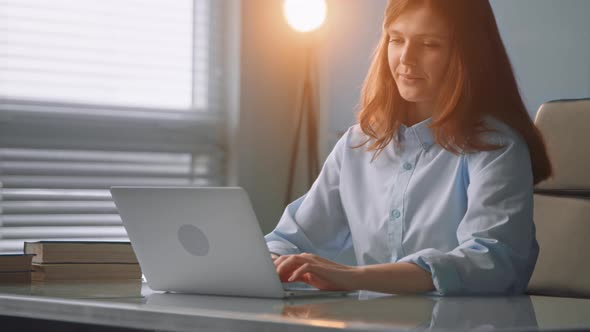 Long red haired woman in blue coat smiles typing on laptop