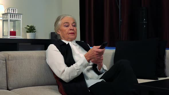 Old Caucasian Woman Goes To Sit on Couch in Living Room and Watches Television