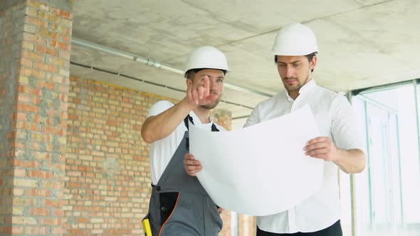 Two Young Engineer in a Safety Helmets Standing at Building Site Holding a Blueprint of the