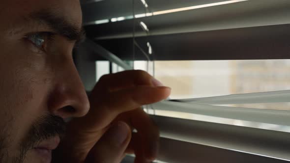 Close-up of a bearded man looking out the window through a slit in the blinds