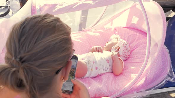 Baby girl in pink bassinet and mother browsing web on cell outdoor