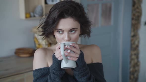 Cute Woman in Cozy Sweater Thoughtfully Drinks Tea in Room