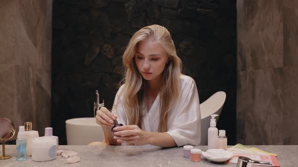 Young Blonde in a White Robe in the Bathroom Applies Cream to Her Face Skincare