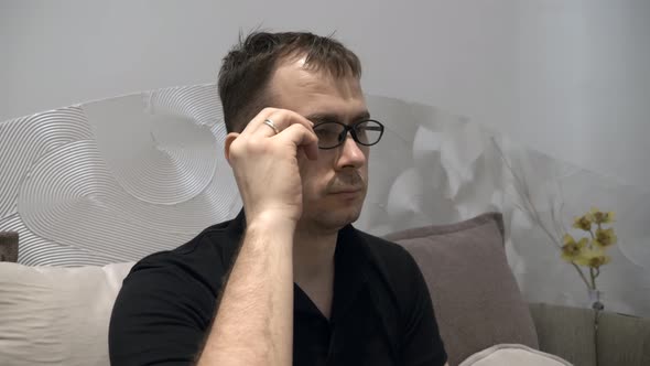 Tired Serious Man in Black Polo Tshirt Sitting on Couch Takes Off His Glasses