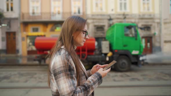 Woman in Glasses and a Coat Walks Down the Street, Uses a Smartphone and Looks Around. Watering