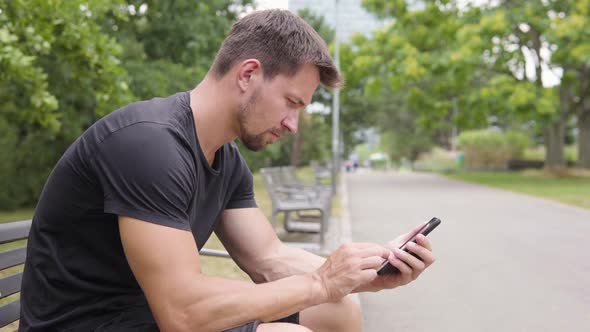 A Young Handsome Man Works on a Smartphone As He Sits on a Bench in a Park - Closeup From the Side