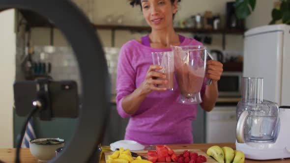 Mixed race female vlogger drinking fruit juice and recording on smartphone in the kitchen at home