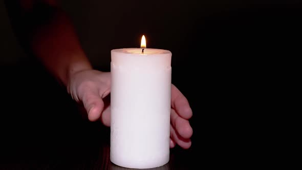 A Female Hand Takes a Burning White Candle From a Table in a Dark Room