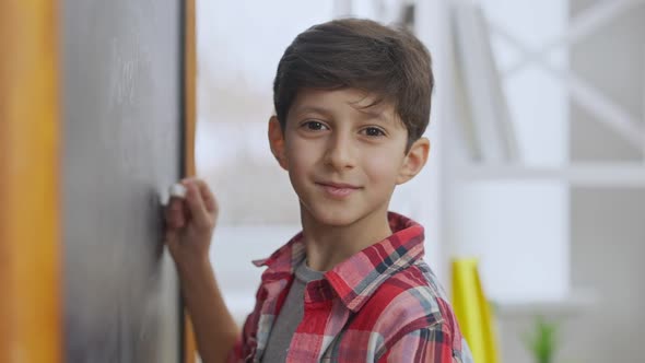 Closeup Portrait of Cute Middle Eastern Schoolboy Writing on Chalkboard Looking at Camera and