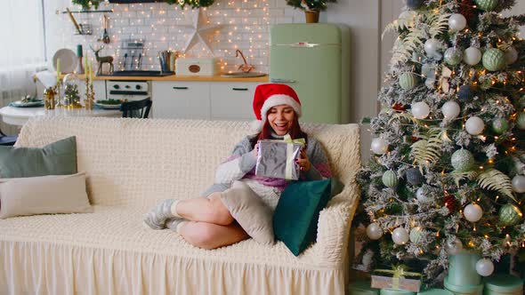 Young Woman in Santa Hat with Gift Box Sitting on Couch Near Christmas Tree in Living Room