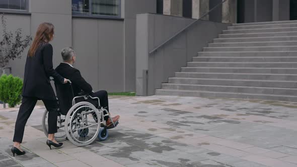 Stylish Woman in Suit Pushing Wheelchair with Man Coworker and Stopping Before Office Building