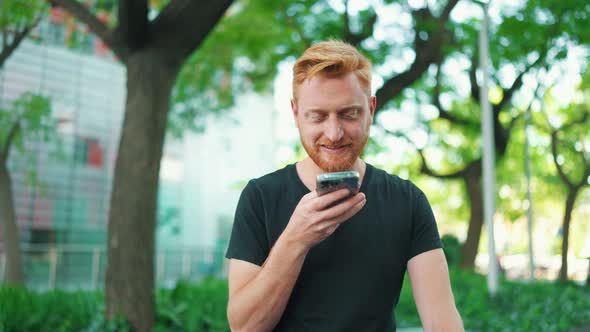 Smiling red haired man recording voice message by phone while sitting on the bench