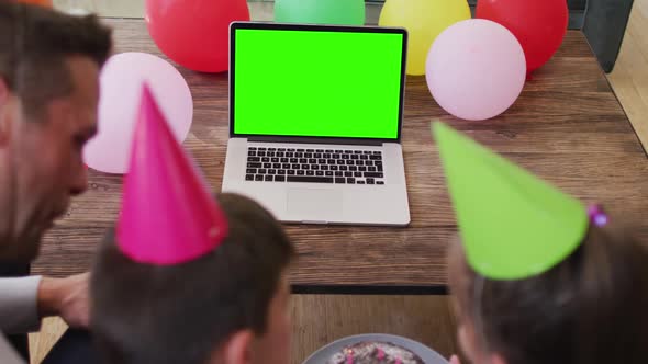 Caucasian family in party hats waving at laptop with green screen on wooden table at home