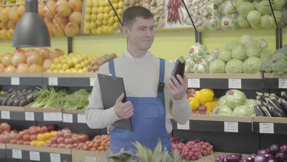 Young Positive Caucasian Employee Posing in Grocery with Folder and Eggplant. Portrait of Smiling