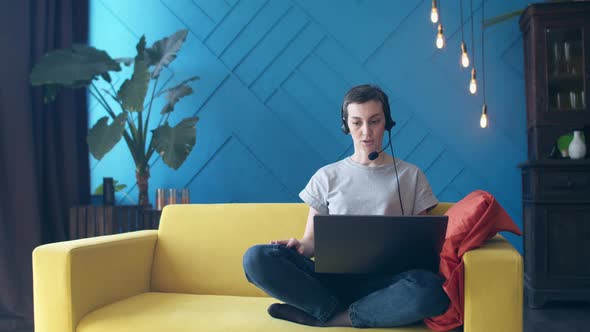 Woman sits on the couch with legs folded and talks through a headset to laptop in room.