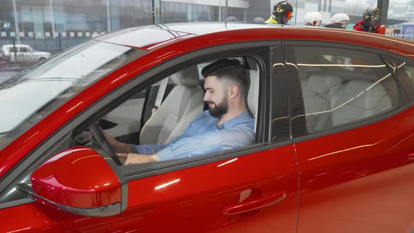 Attractive Young Man Smiling to the Camera Sitting in a New Car at the Dealership