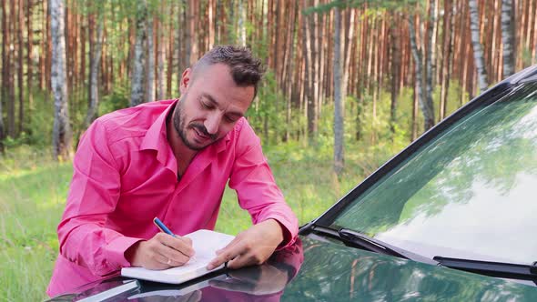 Smiling Businessman Leaning on the Hood Laughs and Makes Notes in a Notebook