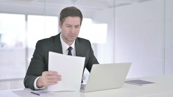 Middle Aged Businessman Reading Documents and Working on Laptop