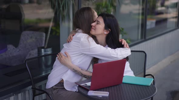 Young Happy Women Hugging in Slow Motion Sitting at Table Outdoors on Cafe Summer Terrace