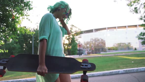 Young Stylish Hipster Girl with Green Hair Walking with Long Board During Sunset or Sunrise Slow