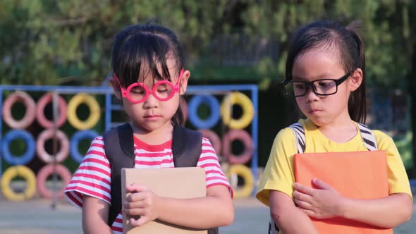 Two cute schoolgirls wearing summer clothes with backpacks walking together in the school alt
