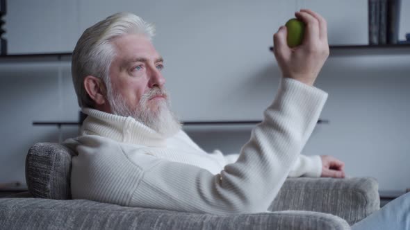 Bearded Elderly Man Sits Alone on a Chair Holding an Apple in His Hand, Thinking