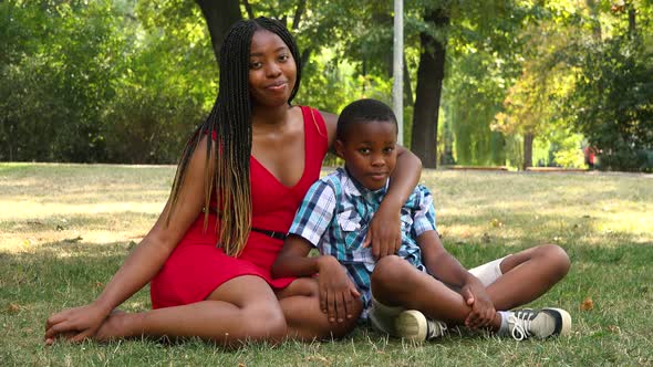 A Young Black Mother and Her Son Sit on Grass in a Park and Smile at the Camera