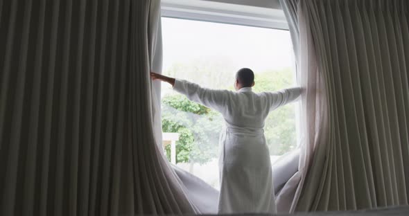 Happy african american plus size woman wearing robe, drawing curtains and looking through window