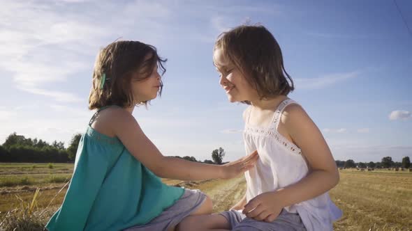 Two Charming Girls Are Sitting on a Roll of Mown Rye in a Field