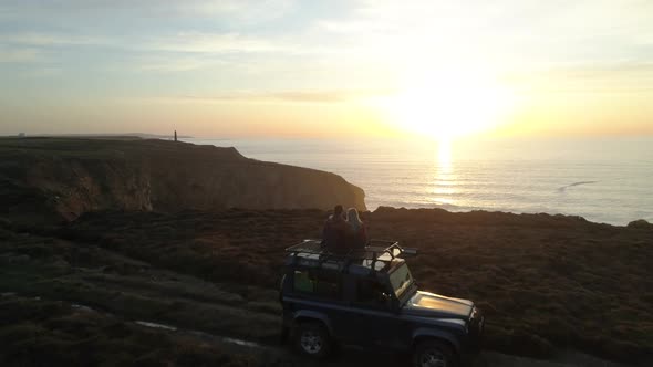 Cinematic aerial shot of cute couple four by four vehicle along ocean cliffs at beautiful sunset