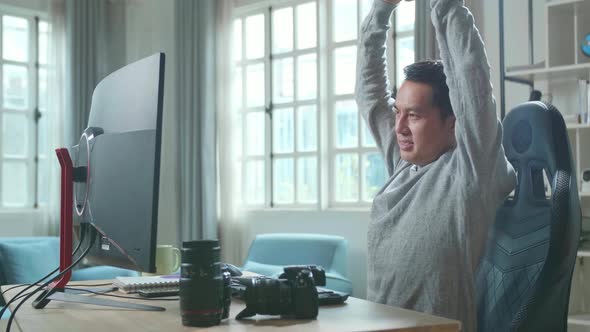 Asian Cameraman Stretching And Leaning Back While Using Desktop Computer For Working At Home