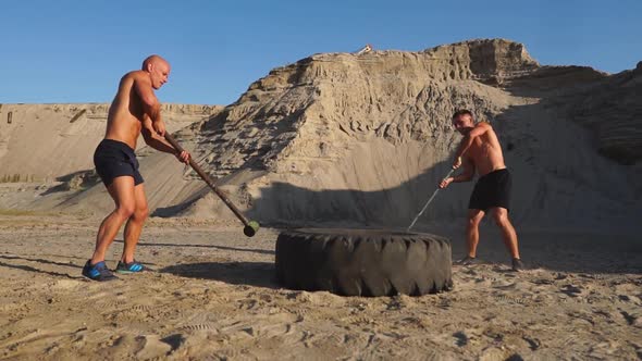 Two Male Athletes Training Together Hit the Wheel with a Hammer at Sunset in the Mountains