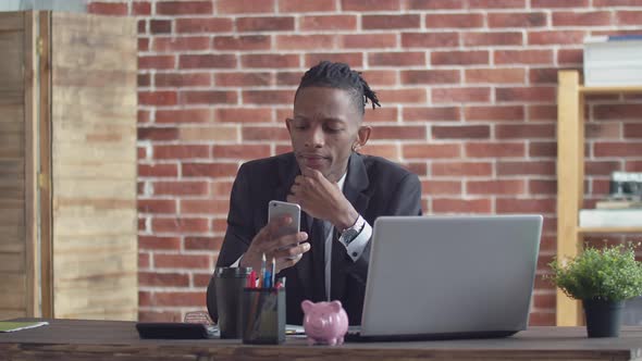 Black Office Man in a Strict Suit Sits at an Office Desk with a Laptop and Uses Phone