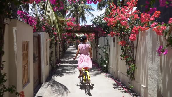 Cute Girl in Lovely Dress Riding Bicycle Through Majestic Tropical Magnificent Passage with Pink