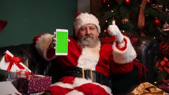 Santa Claus Lies on the Sofa Near a Decorated Christmas Tree and Shows a Tablet with a Green Screen