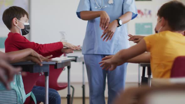 Diverse schoolchildren and medical worker wearing face masks, disinfecting hands in classroom