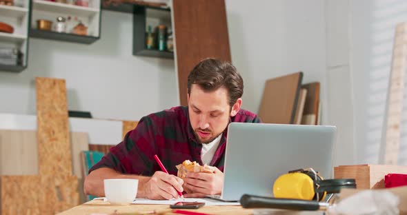 A Handsome Carpenter in a Flannel Shirt Counts the Dimensions on a Laptop in a Carpentry Workshop A