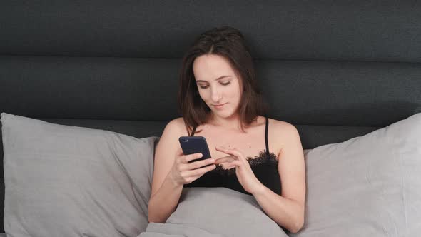 Happy woman typing on smartphone screen on bed at home.