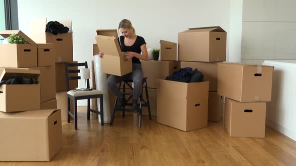 A Moving Woman Sits on a Chair and Puts Things Into a Cardboard Box in an Empty Apartment