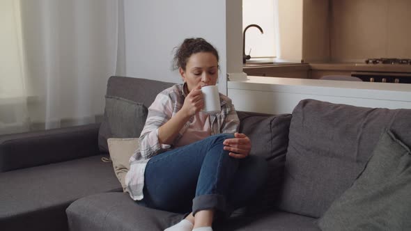 Pensive Adult Woman Sitting on Couch Alone Holding White Mug Indoors