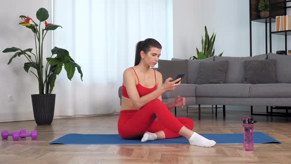 Sportive Beautiful Woman in Sportswear Sitting on Yoga Blue Mat Uses Smartphone