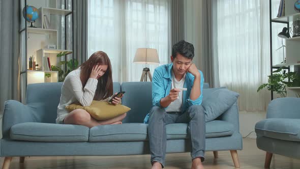 Despaired Man Is Holding Pregnancy Test. Nervous Woman Is Sitting And Messaging On Smartphone