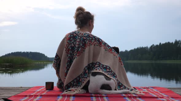 Woman in Cozy Poncho with White and Brown Dog Sitting on the Dock of the Lake at Sunset Enjoying