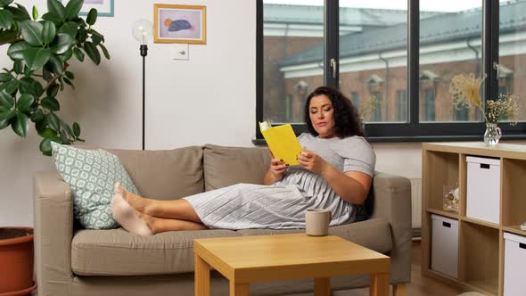 Happy Young Woman Reading Book on Sofa at Home