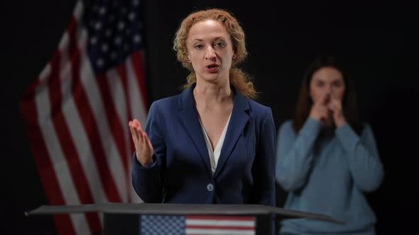 Serious Elegant Politician Woman Talking Looking at Camera with Blurred Sign Language Interpreter